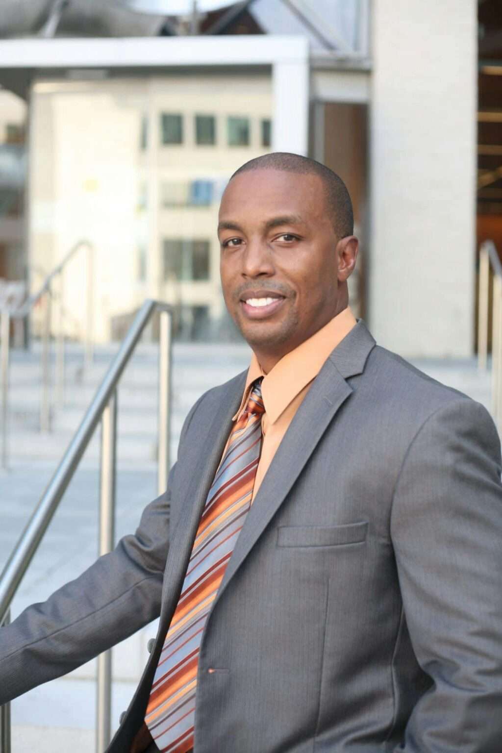 A confident businessman in a suit and tie poses outdoors in a modern urban environment.
